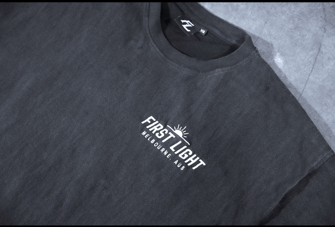Close-up of the embroidered FIRST LIGHT chest detail on the Dawn heavyweight tee, stitched not printed