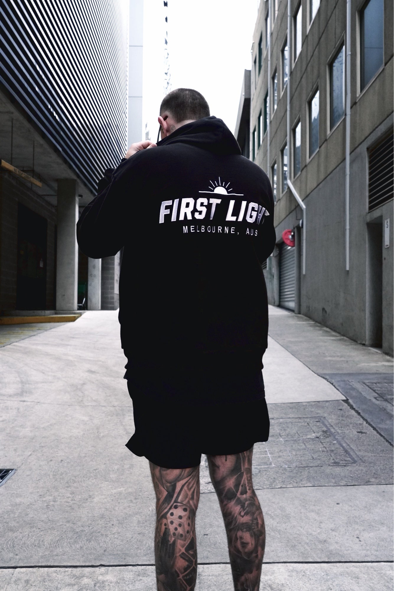 Back view of the Dawn heavyweight hoodie in a Melbourne laneway, embroidered FIRST LIGHT back graphic