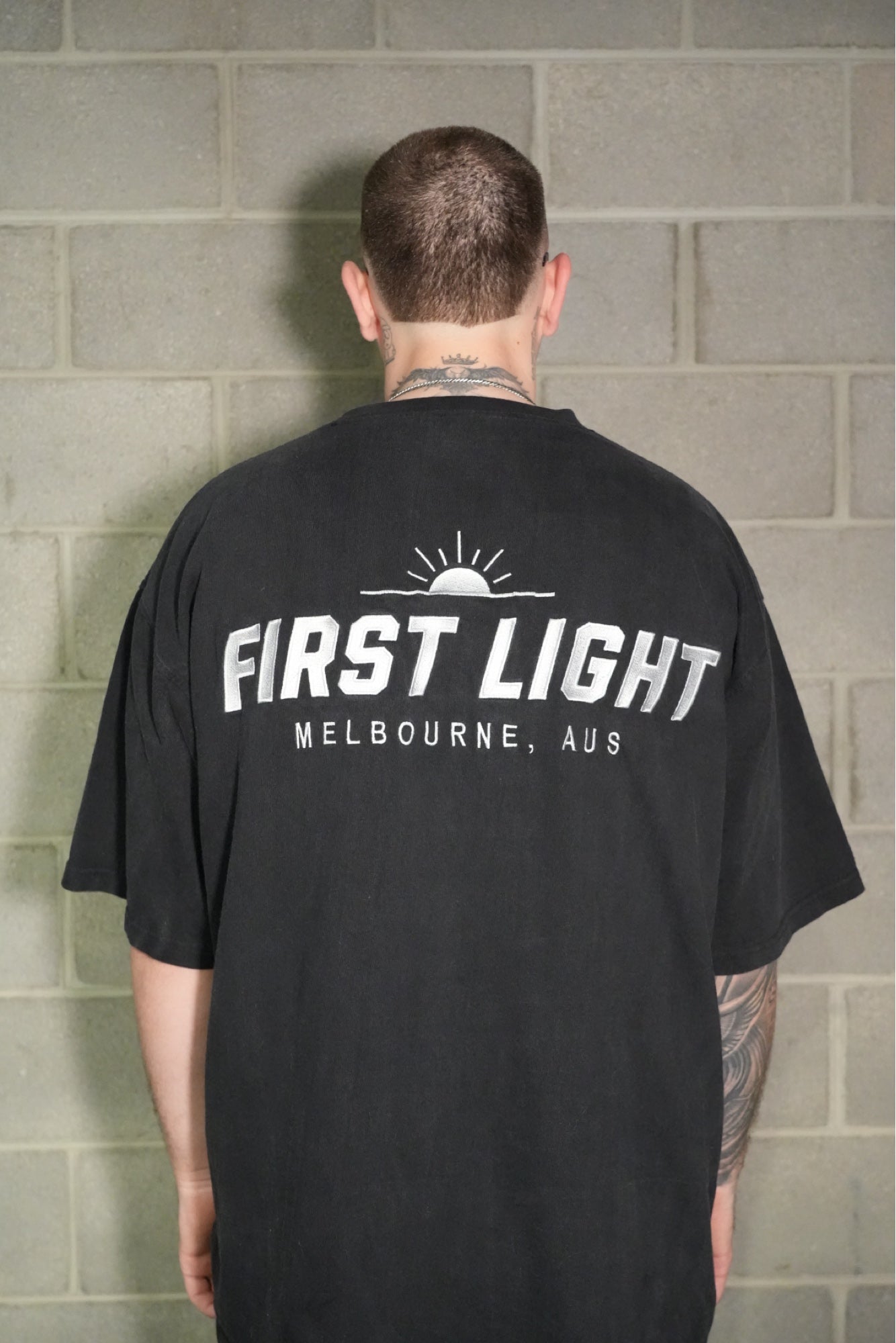 Back view of the FIRST LIGHT Dawn heavyweight tee worn oversized, showing the embroidered FIRST LIGHT Melbourne back graphic